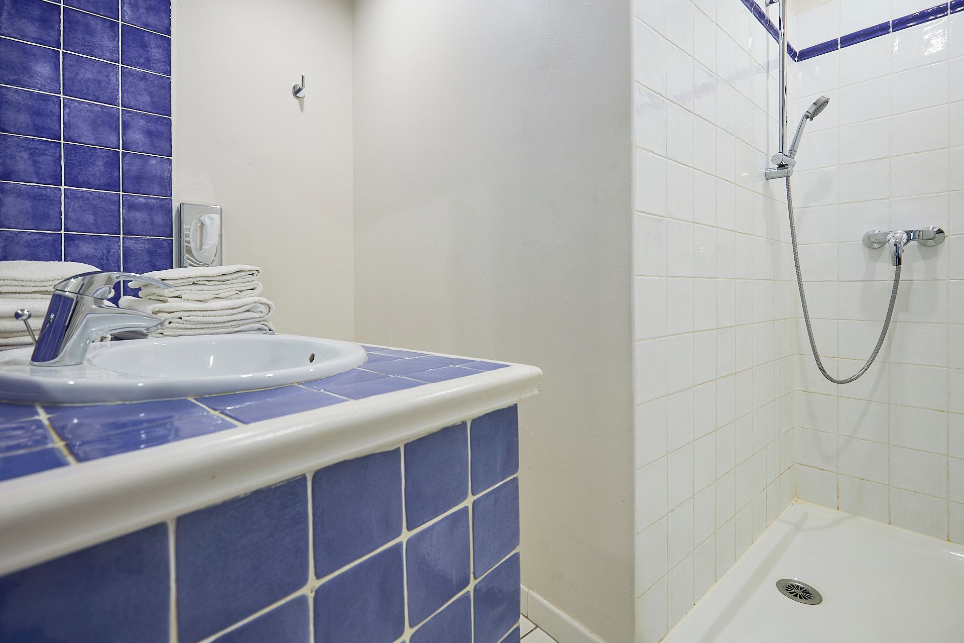 Une salle de bain avec un lavabo, une pile de serviettes blanches, une douche équipée d’un pommeau de douche à main et des murs recouverts de carreaux bleus.