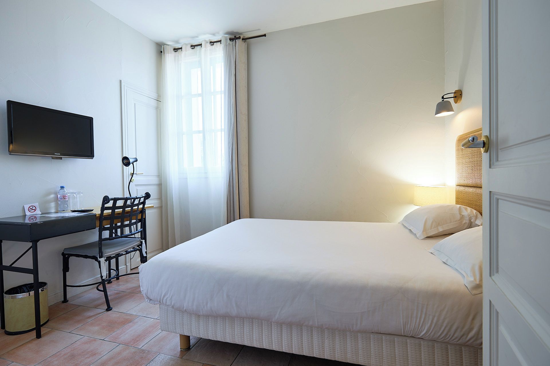 A hotel room with a bed, a desk with a chair, a television, and a wall-mounted lamp.