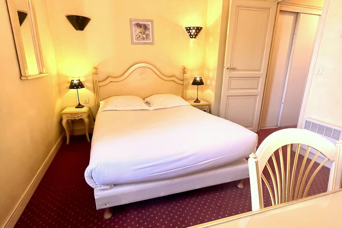Habitación de hotel con una cama grande, dos lámparas de noche, una mesita, una silla y una puerta.