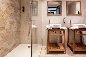 Un baño moderno con cabina de ducha de vidrio, dos lavamanos de madera y un salpicadero de azulejos en patrón de espiga.