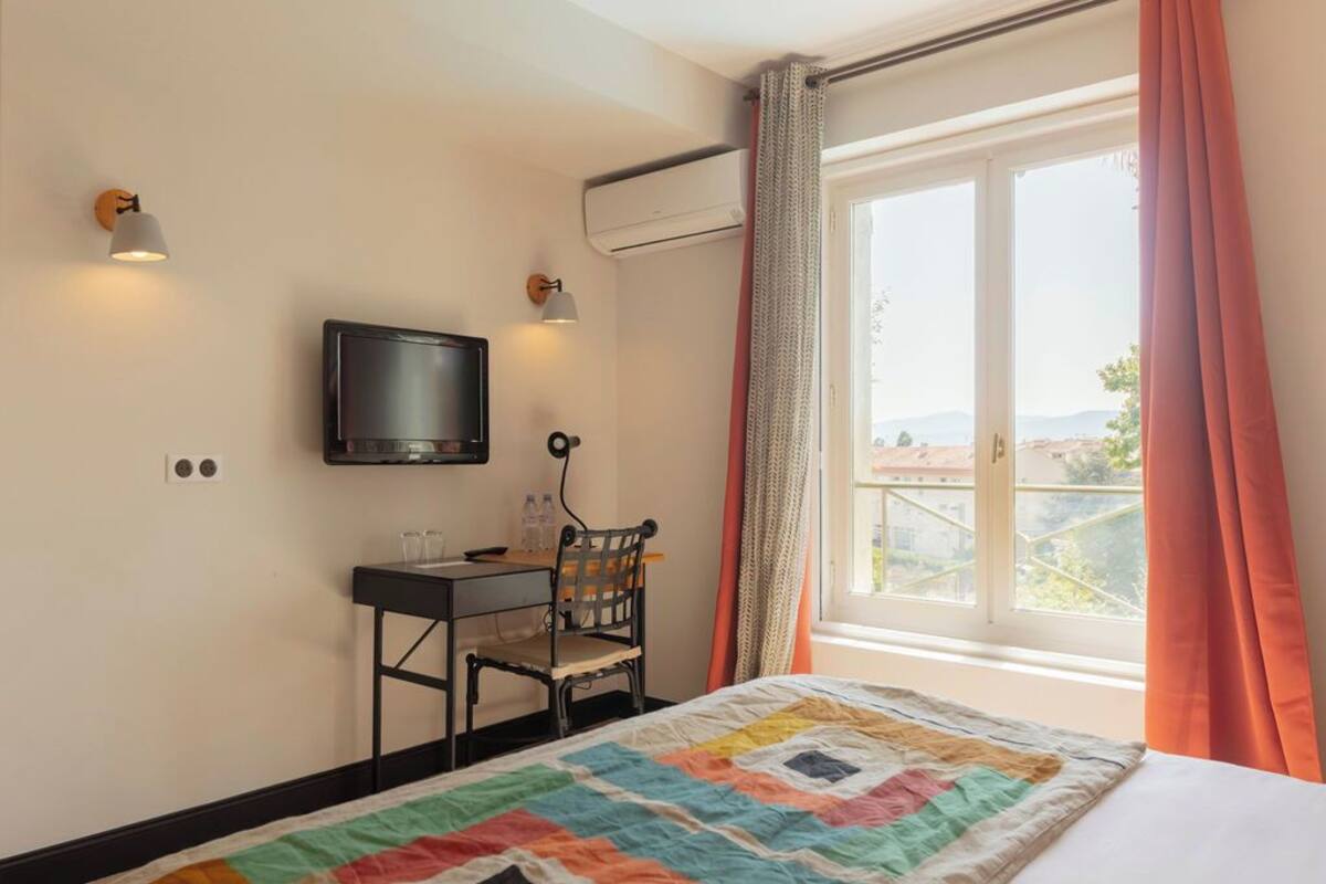 Habitación de hotel con cama, escritorio con silla, televisor y ventana con cortinas.