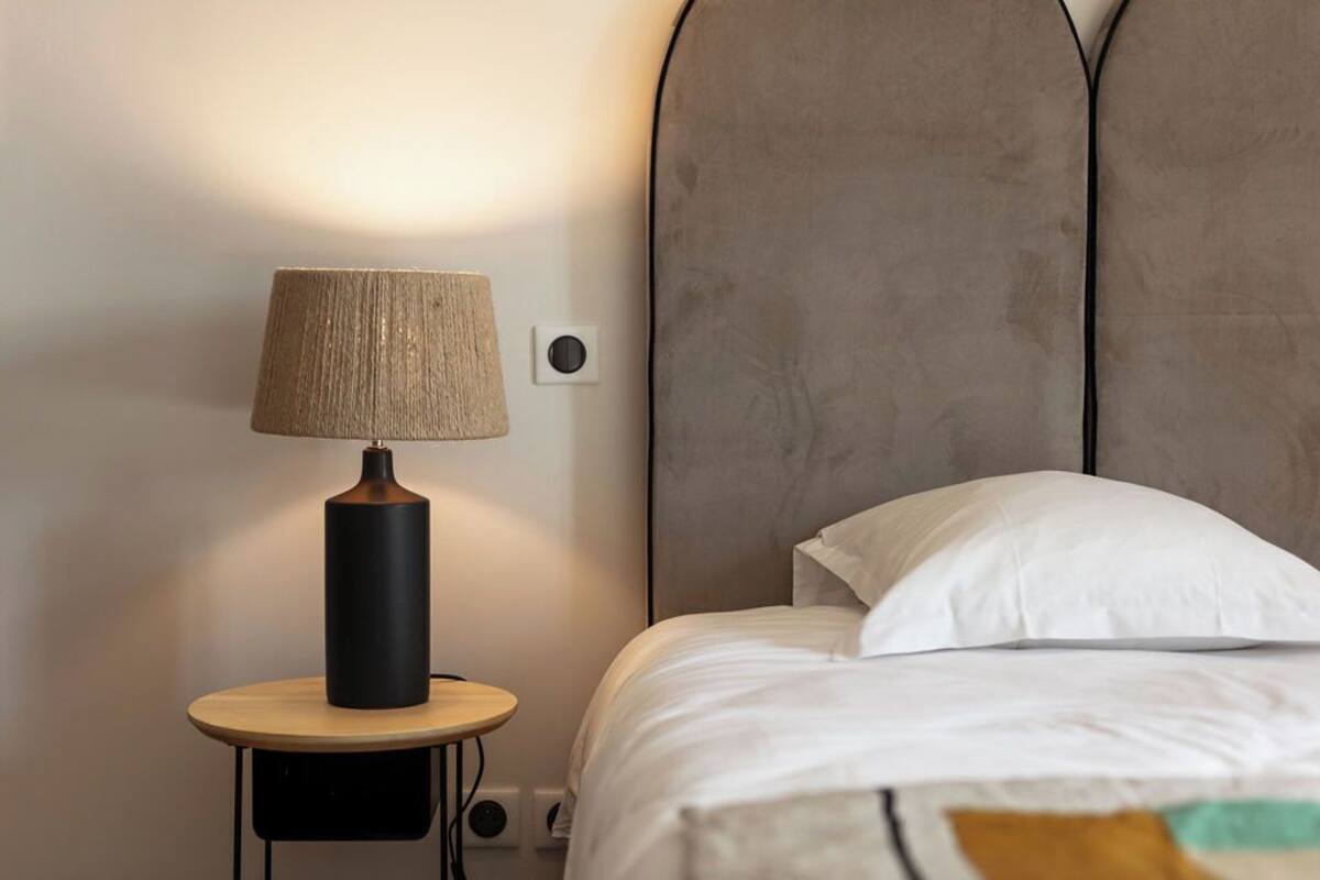 Una cama con ropa de cama blanca, una mesita de noche redonda de madera y un cabecero.