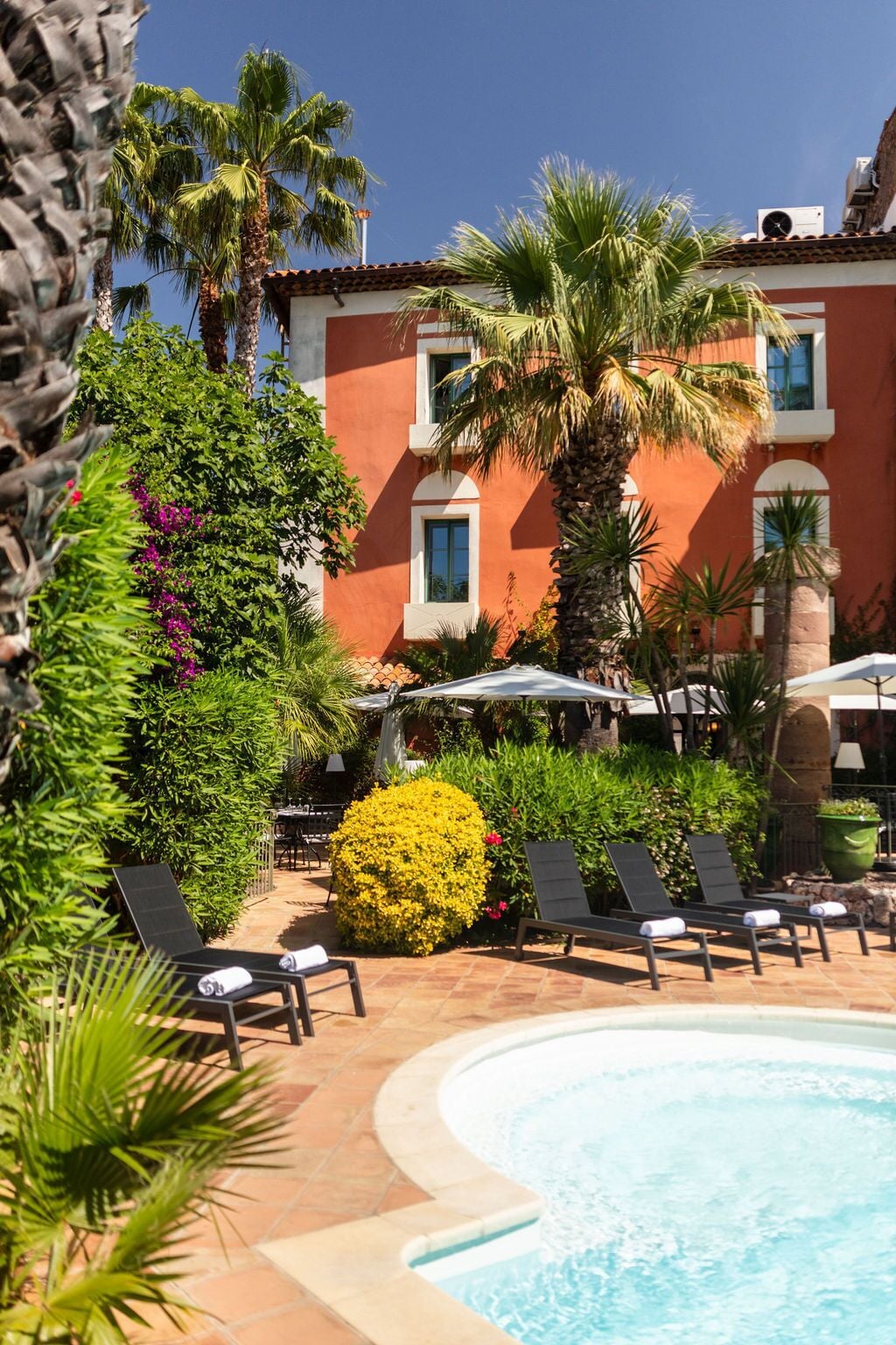 Área de piscina com espreguiçadeiras, uma palmeira e um edifício com paredes vermelhas e janelas.