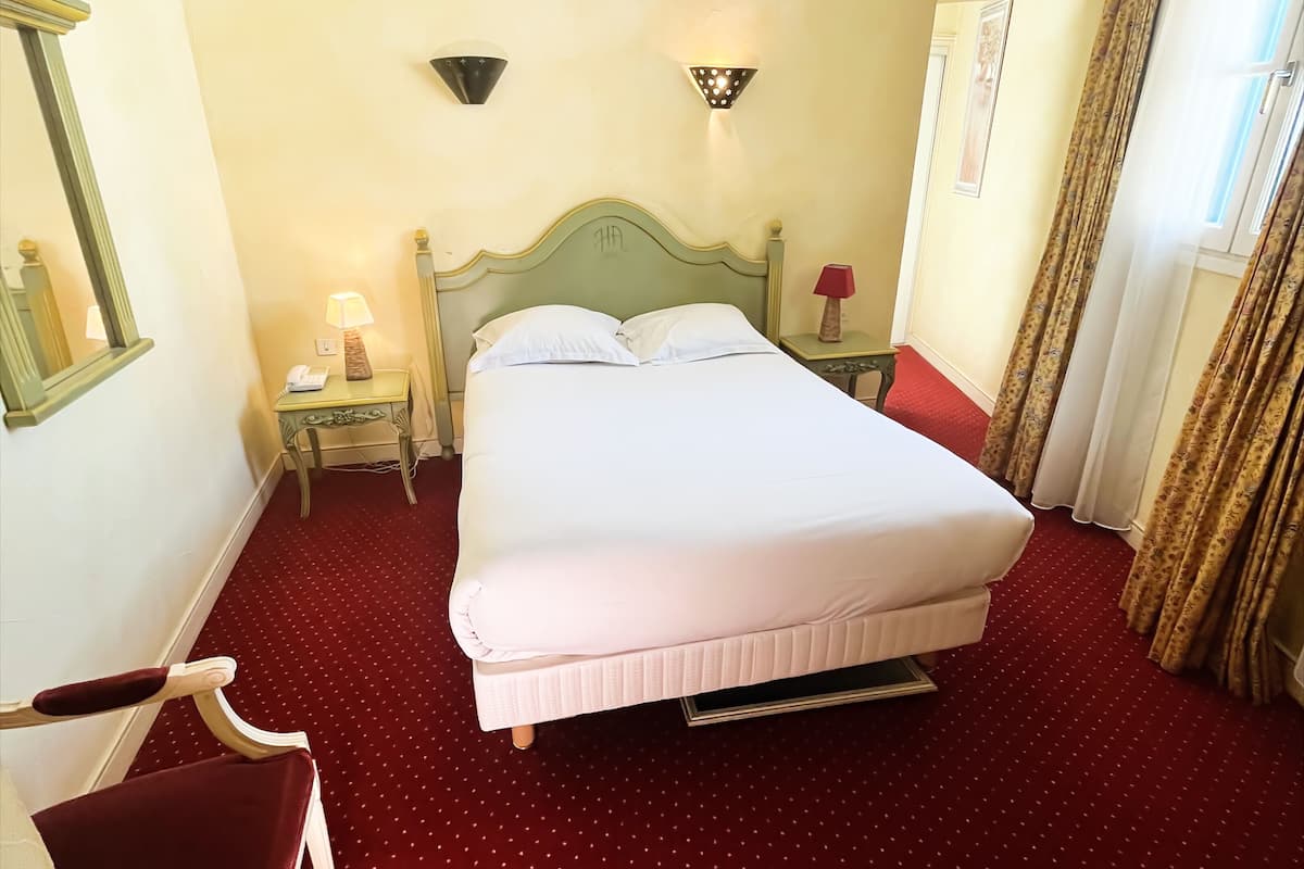 Una cama bien tendida en una habitación con alfombra roja, una mesita de noche con lámpara y un espejo en la pared.