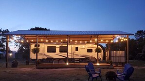 A covered RV with a wooden deck and string lights, two Adirondack chairs, and a fire pit. - "Gone Coastal" Luxury Glamper by the Beach (Carrabelle)