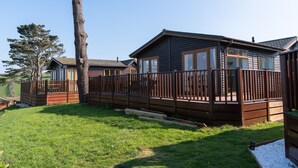 A cabin with a deck, surrounded by a wooden fence and a grassy area. - Serendipity Lodge (Torpoint)
