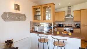 A modern kitchen with wooden cabinets, a built-in microwave, and a coffee maker. There are two bar stools and a sign that reads "Be our guest." - Serendipity Lodge (Torpoint)