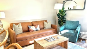 Una sala de estar con un sofá de cuero marrón, un sillón verde azulado, una mesa de centro de madera y una planta grande de hojas verdes.