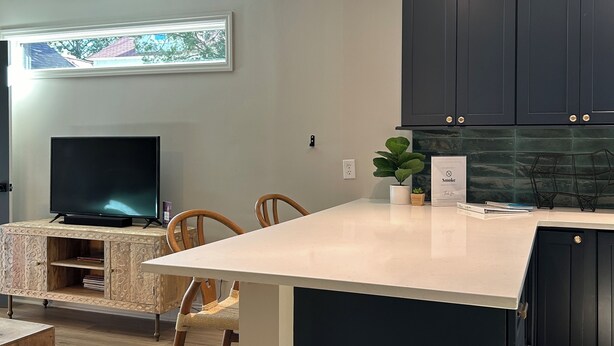 Una cocina moderna con armarios oscuros, encimera blanca, televisor integrado y un comedor con sillas.