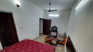 A room with a bed, a small table, a sofa, a ceiling fan, and a television on a stand. - Janardan Homestay Lucknow (Lucknow)