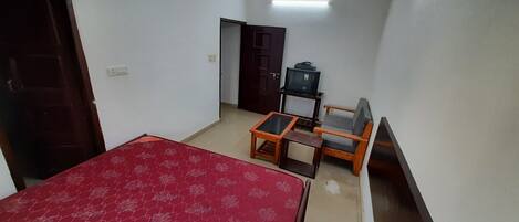 Quarto com cama, mesa pequena, sofá, ventilador de teto e televisão sobre um suporte.