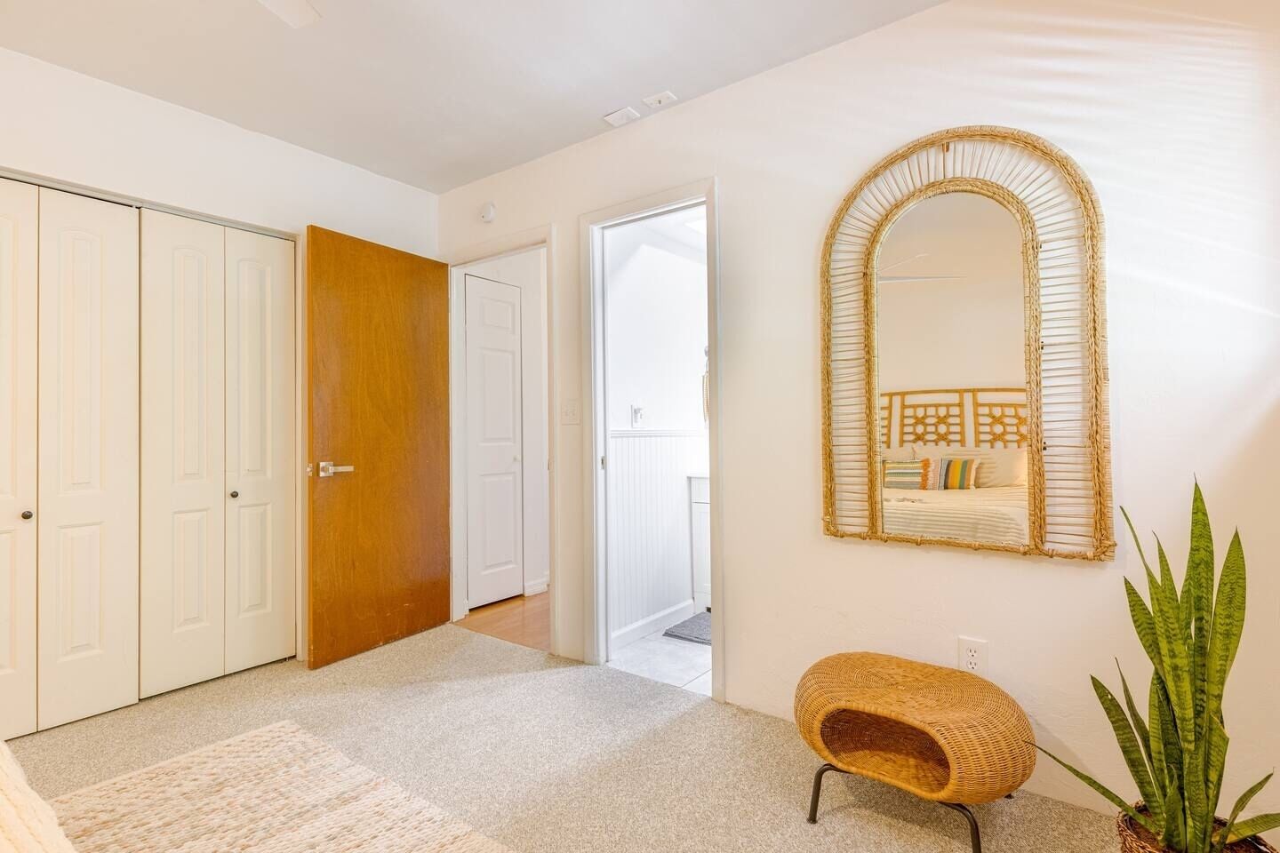 A hallway with white doors, a wooden door, a woven chair, a mirror, and a potted plant.