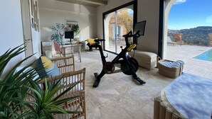 A spacious living area with a large window, a stationary exercise bike, a dining table, and a view of a pool and landscape. - Splendid 6-bedroom villa with sea view in Grasse (Grasse)