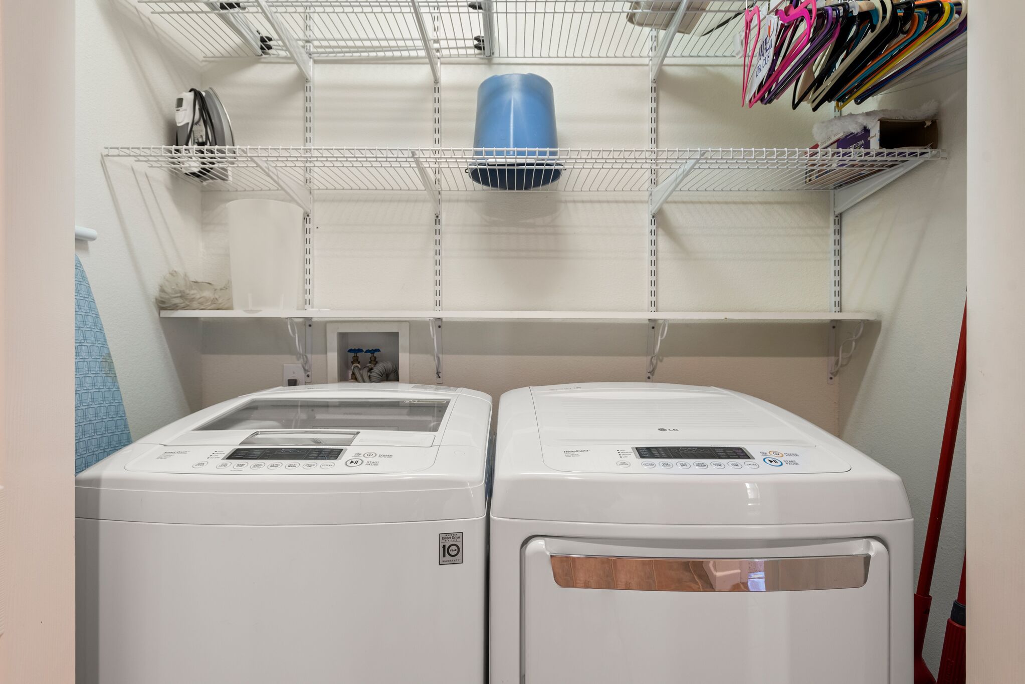 A laundry room with two white front-loading washing machines, a blue vent, and shelves above holding various items.