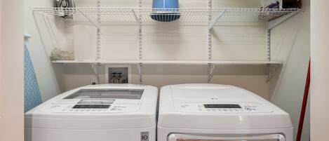 A laundry room with two white front-loading washing machines, a blue vent, and shelves above holding various items.