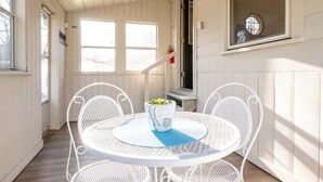 Une table en métal blanc, accompagnée de quatre chaises et d’une plante en pot, est placée sur une véranda aux murs en bois et dotée d’une fenêtre.