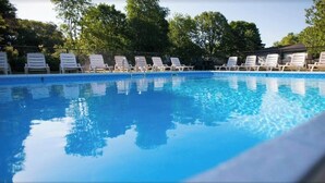 Une piscine bordée de chaises longues blanches, alignées le long de son pourtour, et entourée d’arbres.