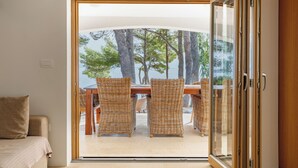 Habitación con puerta corrediza de vidrio que da a un espacio al aire libre con mobiliario de mimbre y vistas a árboles y un cuerpo de agua.