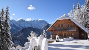 A wooden cabin with a balcony, surrounded by snow-covered trees and mountains in the background.