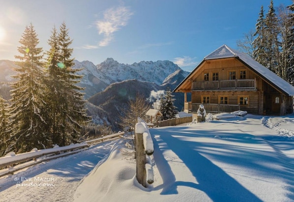 A wooden cabin with a balcony, surrounded by snow-covered mountains and trees.