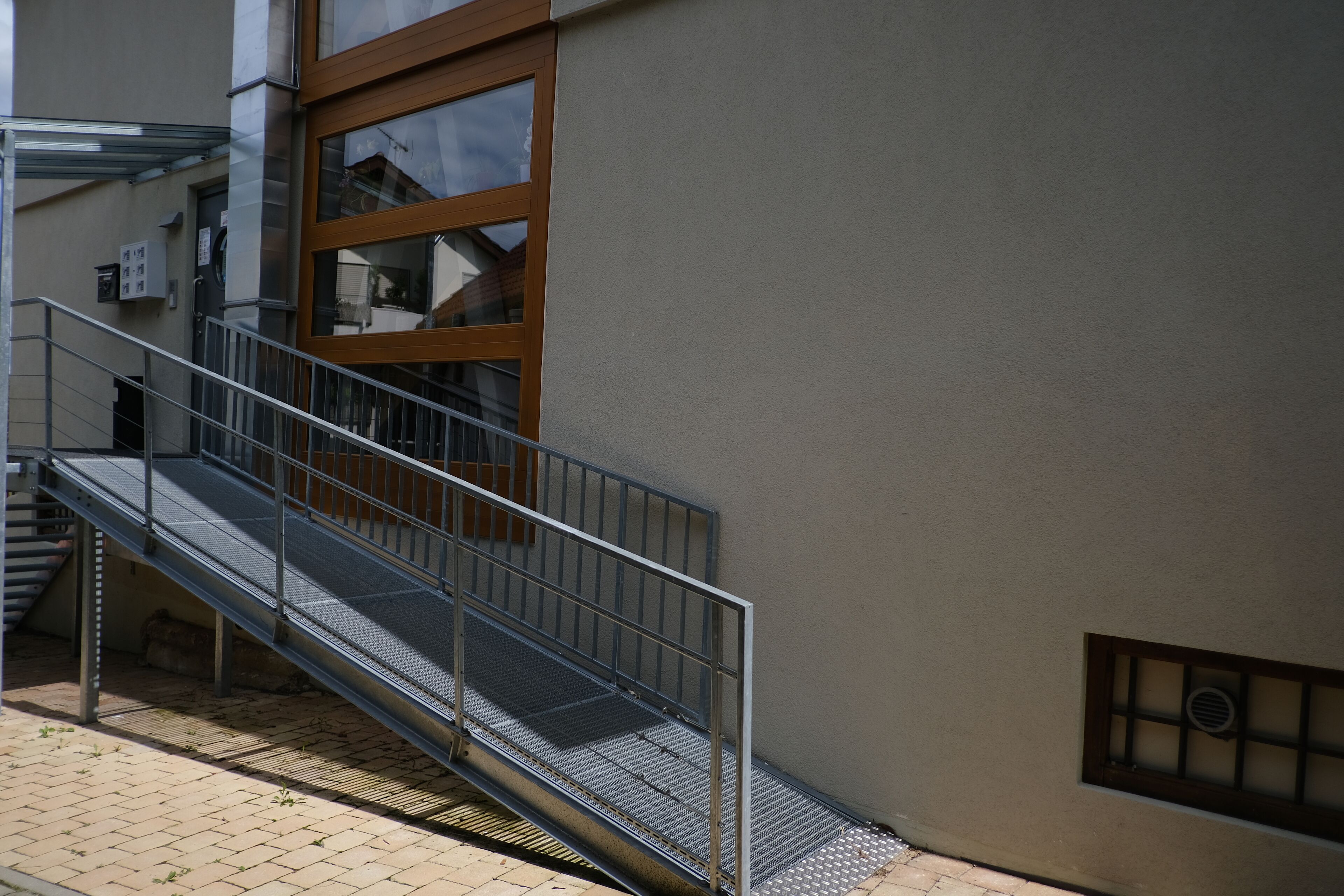 Una escalera metálica conduce a la entrada de un edificio con una puerta de cristal y una rejilla en la pared.