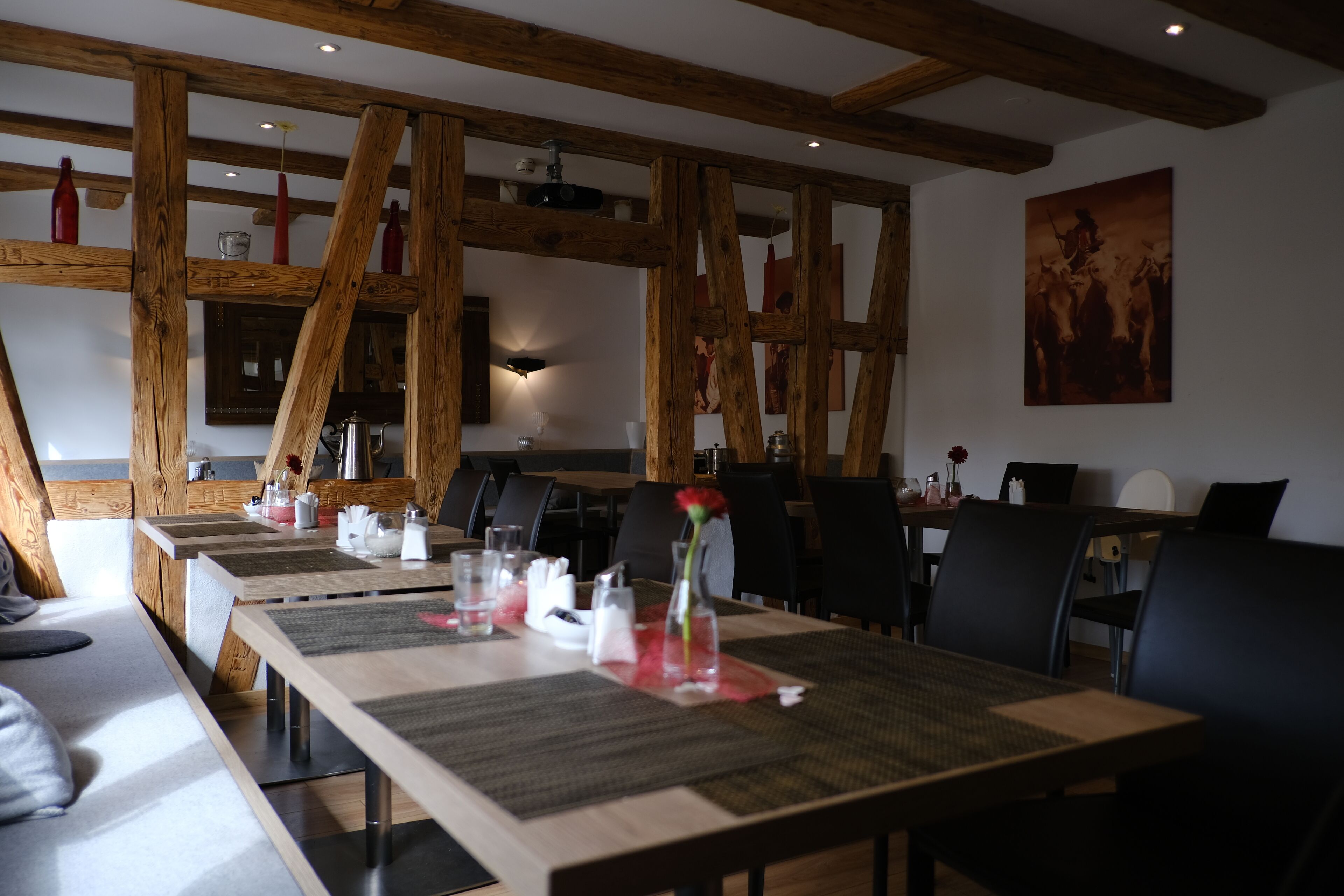 Un comedor con vigas de madera, mesas dispuestas con individuales y un jarrón con una flor roja.