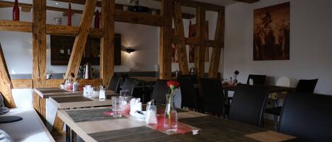 Un comedor con vigas de madera, mesas dispuestas con individuales y un jarrón con una flor roja.