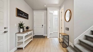 A hallway with a wooden floor, a white console table with a plant, a black sign saying "HELLO AND welcome", and a staircase with a grey carpet. - Paseos on the Green, PRIVATE HOT TUB, PUTTING GREEN! (Washington)