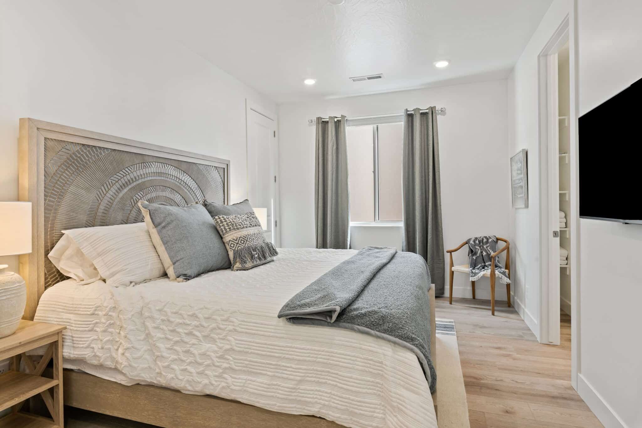 A bedroom with a large bed, a bedside table, a chair, a window with curtains, and a TV on the wall.