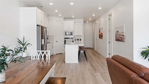 A modern kitchen with white cabinetry, a central island, and a dining area with a wooden table and benches. - Cloud 9 At Desert Color: Desert Color Lagoon + Beaches (St. George)