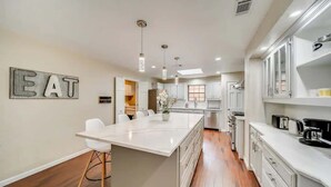 A modern kitchen with white cabinetry, a central island, pendant lighting, and a 'EAT' sign on the wall. - Stunning Gables House, Basketball Court (Dallas)