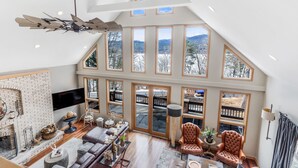 A spacious loft-style living area with a high ceiling, large windows, a fireplace, and a ceiling fan. - Quaint + Modern Lakeview Home on Sacandaga Lake (Hadley)