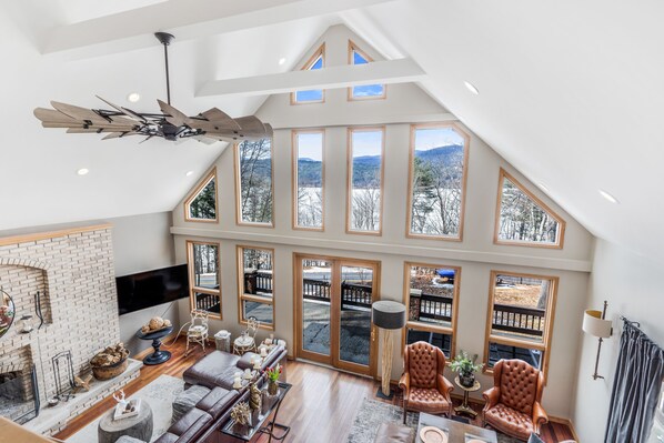A spacious loft-style living area with a high ceiling, large windows, a fireplace, and a ceiling fan.