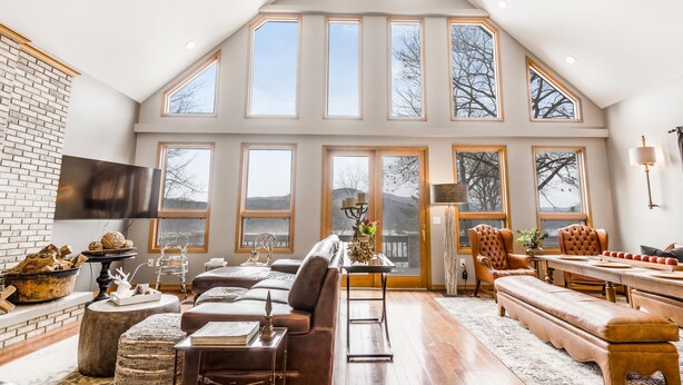 A spacious living room with a high ceiling, large windows, a fireplace, and a dining area. - Quaint + Modern Lakeview Home on Sacandaga Lake (Hadley)