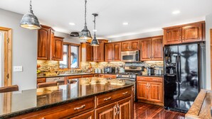 A modern kitchen with wooden cabinets, granite countertops, stainless steel appliances, and pendant lighting. - Quaint + Modern Lakeview Home on Sacandaga Lake (Hadley)