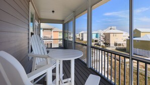 Une véranda couverte, avec des chaises blanches et une table, donnant sur un quartier résidentiel avec des maisons et une plage.