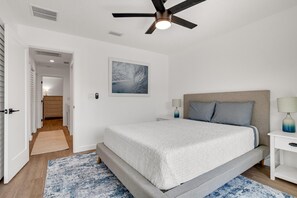 Un dormitorio con cama, ventilador de techo, mesita de noche, lámpara y un cuadro en la pared.