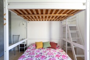 Uma cama beliche com teto de madeira e escada, uma colcha florida e uma pequena mesa com gavetas.