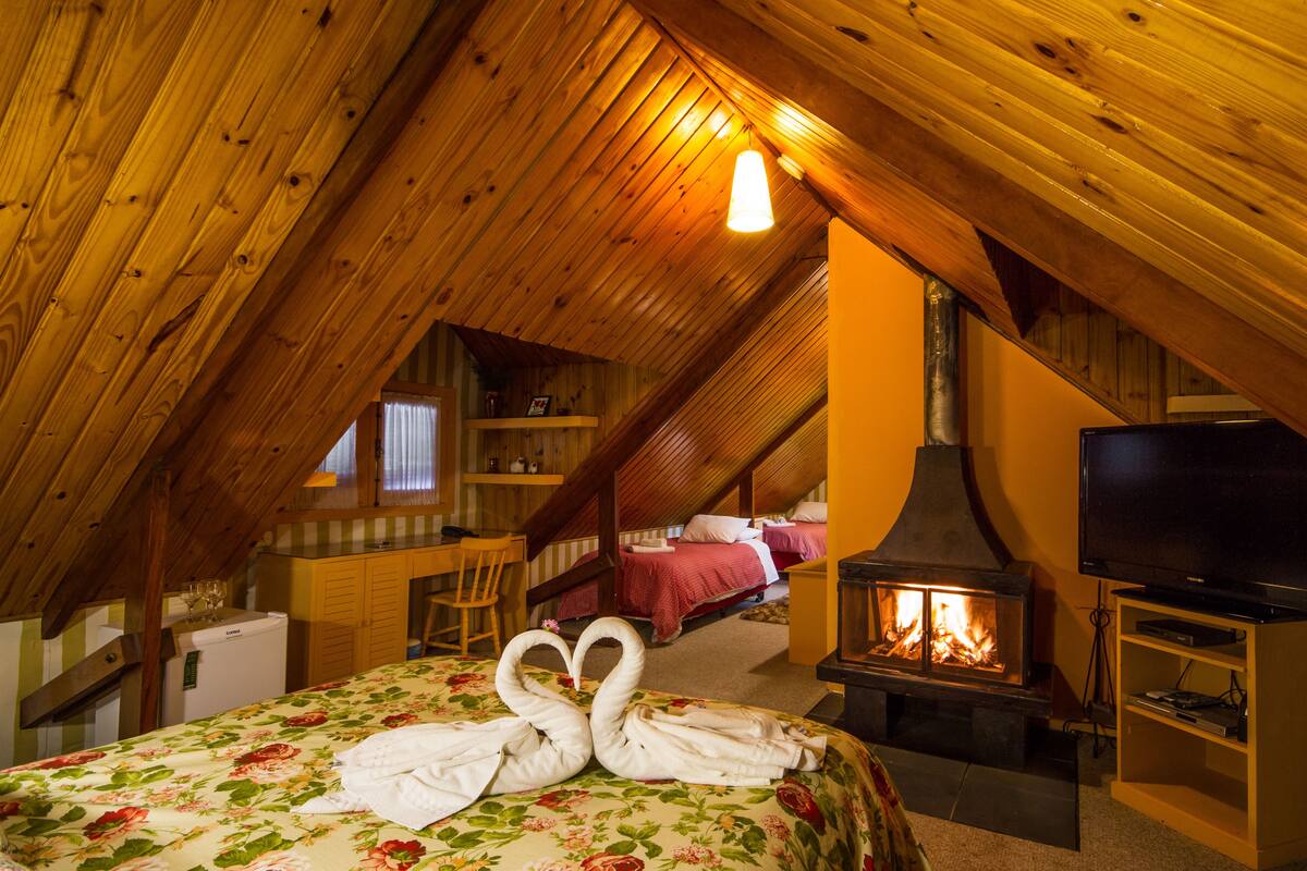 Ein gemütliches Schlafzimmer im Dachgeschoss mit Holzbalkendecke, Kamin, Fernseher und einem Bett mit zu Schwänen geformten Handtüchern.