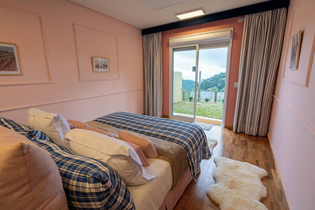Ein Schlafzimmer mit zwei Betten, einem großen Fenster mit Vorhängen, Blick ins Grüne und einem weißen Fellteppich auf dem Boden.