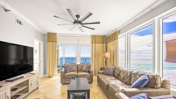 A spacious living room with a large flat-screen TV, leather sofas, a coffee table, and a ceiling fan. - Jade East Towers~Beach Front w/Amazing Unparalleled  Sunrise and Sunset Views (Jade East Towers , Destin)