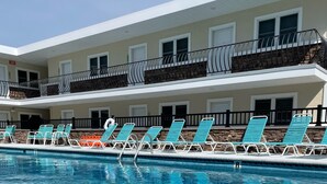 A swimming pool with clear blue water, surrounded by lounge chairs and a building with balconies. - Beach block Wildwood Crest ground level condo-103 (Wildwood Crest)