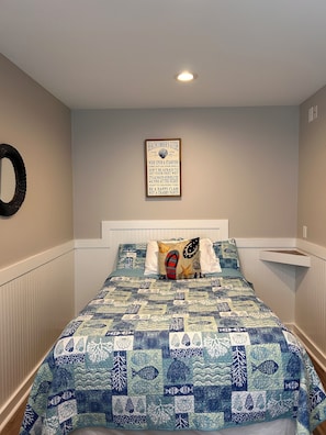 A bedroom with a bed featuring a blue and white quilt, a wall-mounted decorative sign, and a white headboard.