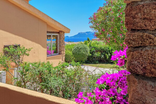 A Mediterranean-style building with a stone facade, a balcony with a stone balustrade, and a view of the sea and mountains in the background.