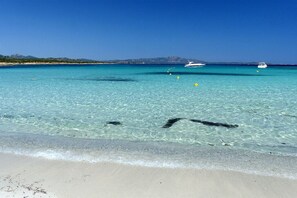 Une plage à l’eau turquoise et limpide, un rivage sablonneux et des bateaux à l’horizon.