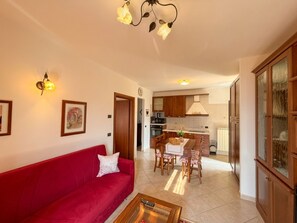 Un salon avec un canapé rouge, une table basse et un coin repas comprenant une table et des chaises. Au fond, on aperçoit une cuisine avec des meubles en bois et un fourneau.