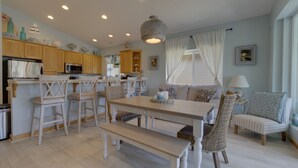 Une cuisine chaleureuse et bien éclairée, avec des meubles en bois, un coin repas comprenant une table et des chaises, et un espace détente avec un canapé et des coussins.