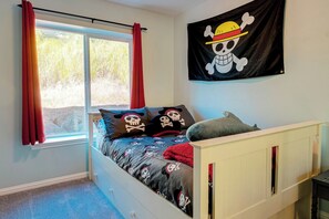 A bedroom with a bed, a window with curtains, a wall-mounted pirate-themed flag, and a small table. - Friends & Family Seaside Retreat, Walk to Beach! (Gold Beach)