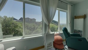Une pièce dotée de grandes fenêtres, d’un fauteuil turquoise, d’un petit pouf orange et d’une bibliothèque remplie de livres.