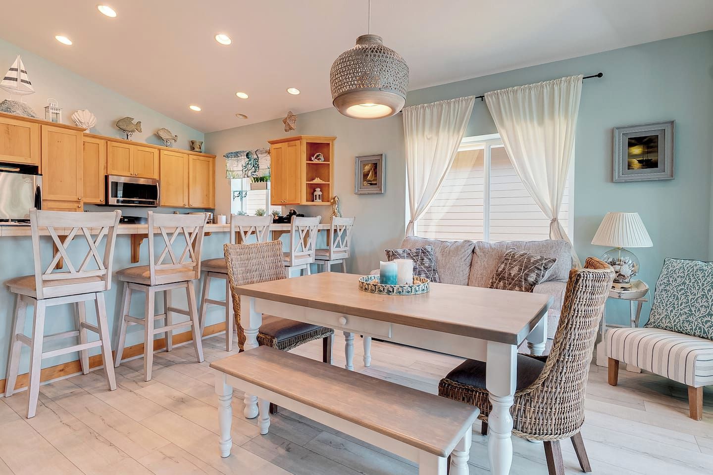 A dining area with a wooden table and chairs, a hanging light fixture, and a view of the kitchen in the background.
